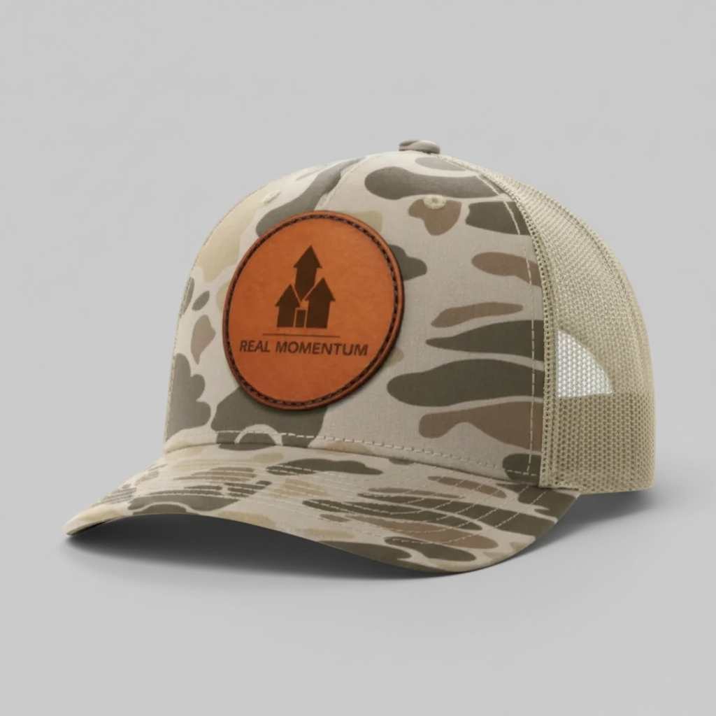 Camouflage trucker hat with a brown leather patch featuring 'Real Momentum' logo on a white background.
